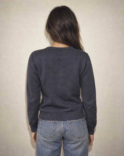 A woman viewed from behind wearing a dark charcoal gray knit sweater and light blue jeans, standing against a textured beige wall.