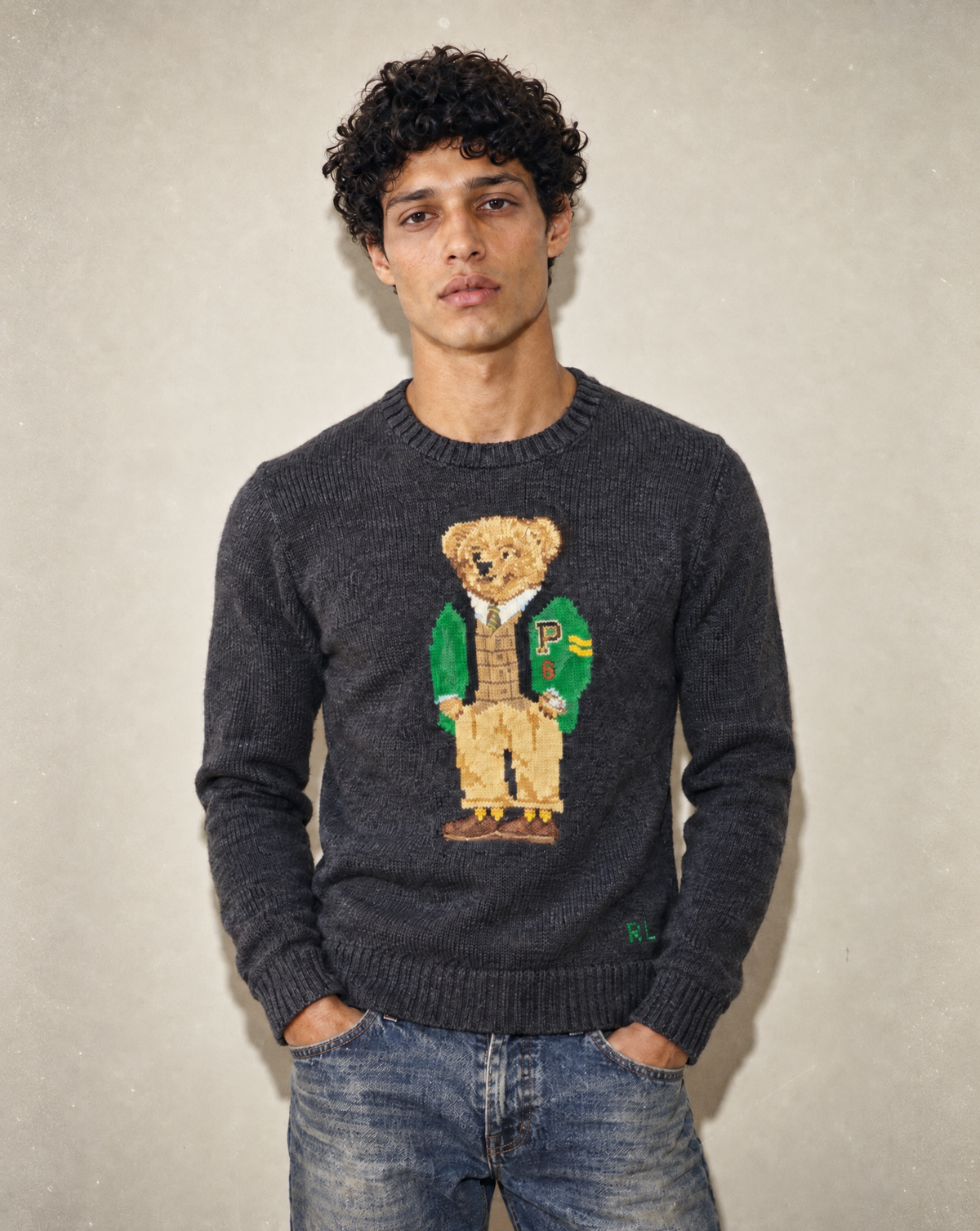 A man wearing a dark gray knit sweater with a large embroidered teddy bear graphic on the chest, paired with blue jeans, standing against a textured light gray wall.