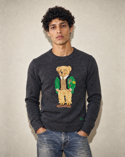 A man wearing a dark gray knit sweater with a large embroidered teddy bear graphic on the chest, paired with blue jeans, standing against a textured light gray wall.