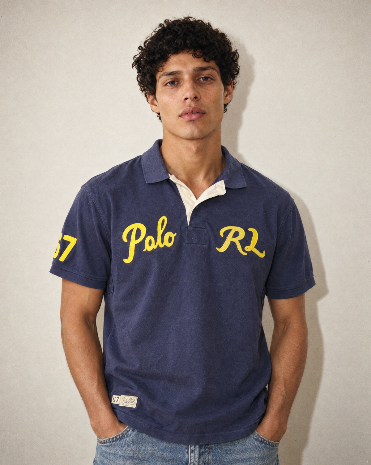 A man wearing a navy blue Polo Ralph Lauren polo shirt with yellow embroidered lettering on the chest and sleeve, paired with blue jeans, standing against a neutral light gray wall.