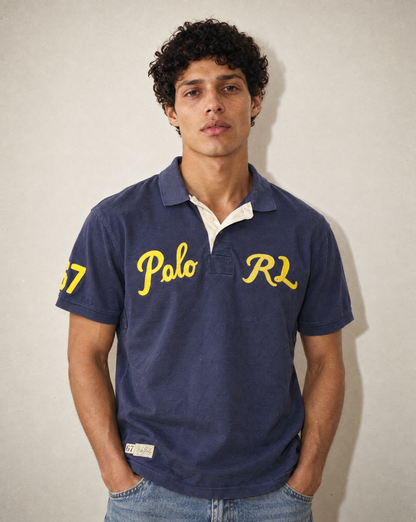A man wearing a navy blue Polo Ralph Lauren polo shirt with yellow embroidered lettering on the chest and sleeve, paired with blue jeans, standing against a neutral light gray wall.