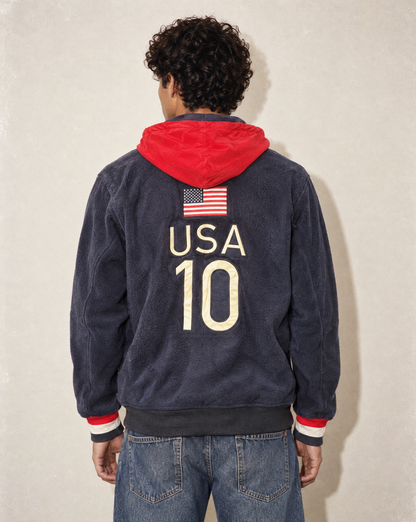 A person shown from the back wearing a navy hooded jacket with a bright red hood, large cream 'USA 10' lettering, and a small American flag graphic on the upper back, paired with blue jeans against a light textured wall.