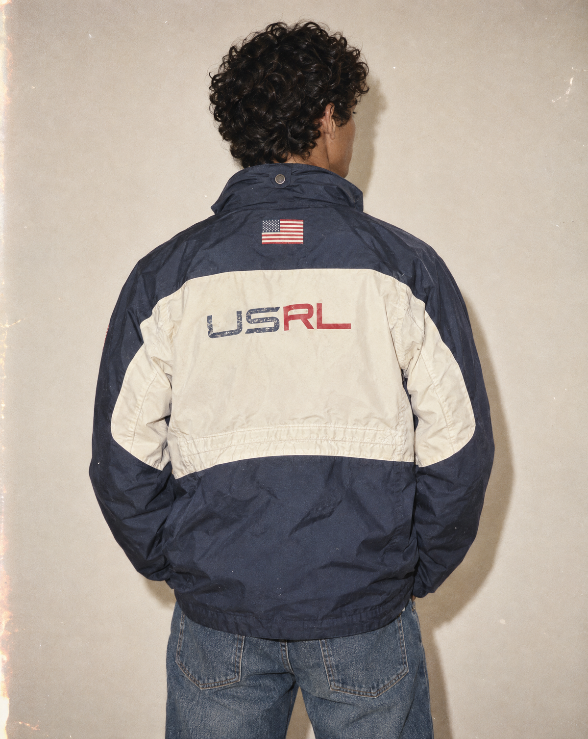 A person viewed from behind wearing a colorblock windbreaker jacket with navy blue and off-white panels, featuring 'USRL' lettering and an American flag patch on the back, paired with blue jeans against a textured beige wall.