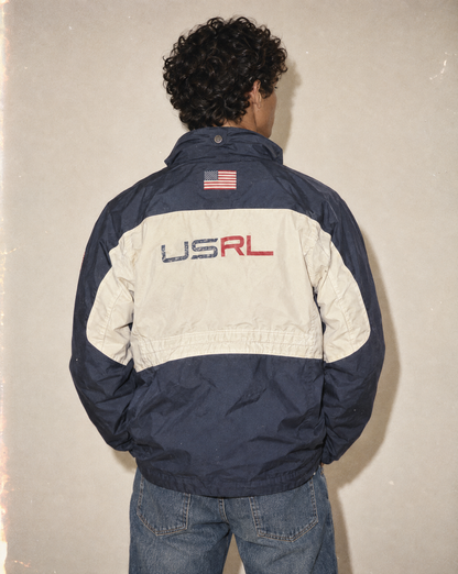 A person viewed from behind wearing a colorblock windbreaker jacket with navy blue and off-white panels, featuring 'USRL' lettering and an American flag patch on the back, paired with blue jeans against a textured beige wall.
