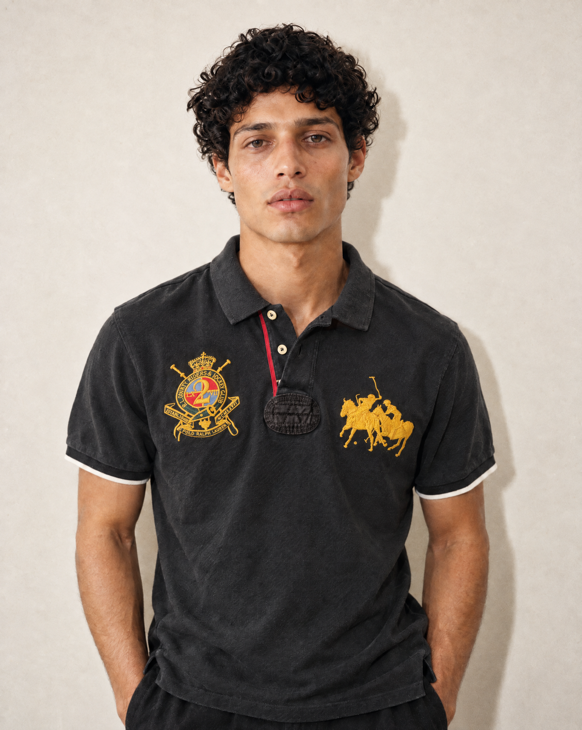 A man wearing a black Polo Ralph Lauren polo shirt with gold embroidered logos on the chest, standing against a neutral beige wall.