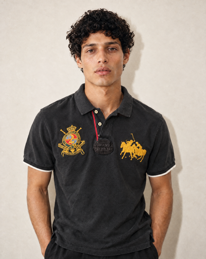 A man wearing a black Polo Ralph Lauren polo shirt with gold embroidered logos on the chest, standing against a neutral beige wall.
