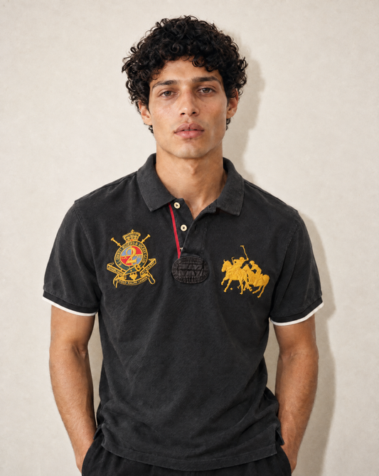 A man wearing a black Polo Ralph Lauren polo shirt with gold embroidered logos on the chest, standing against a neutral beige wall.