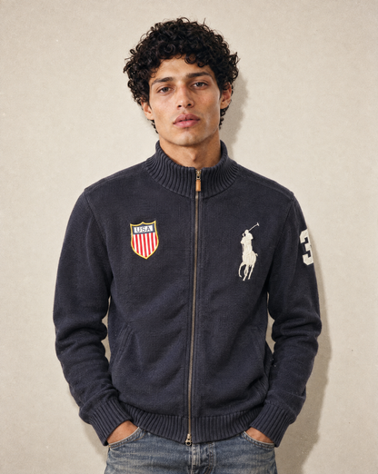 A man wearing a dark navy blue zip-up sweater with the Polo Ralph Lauren logo and a shield crest on the chest, paired with blue jeans, standing against a neutral beige wall.