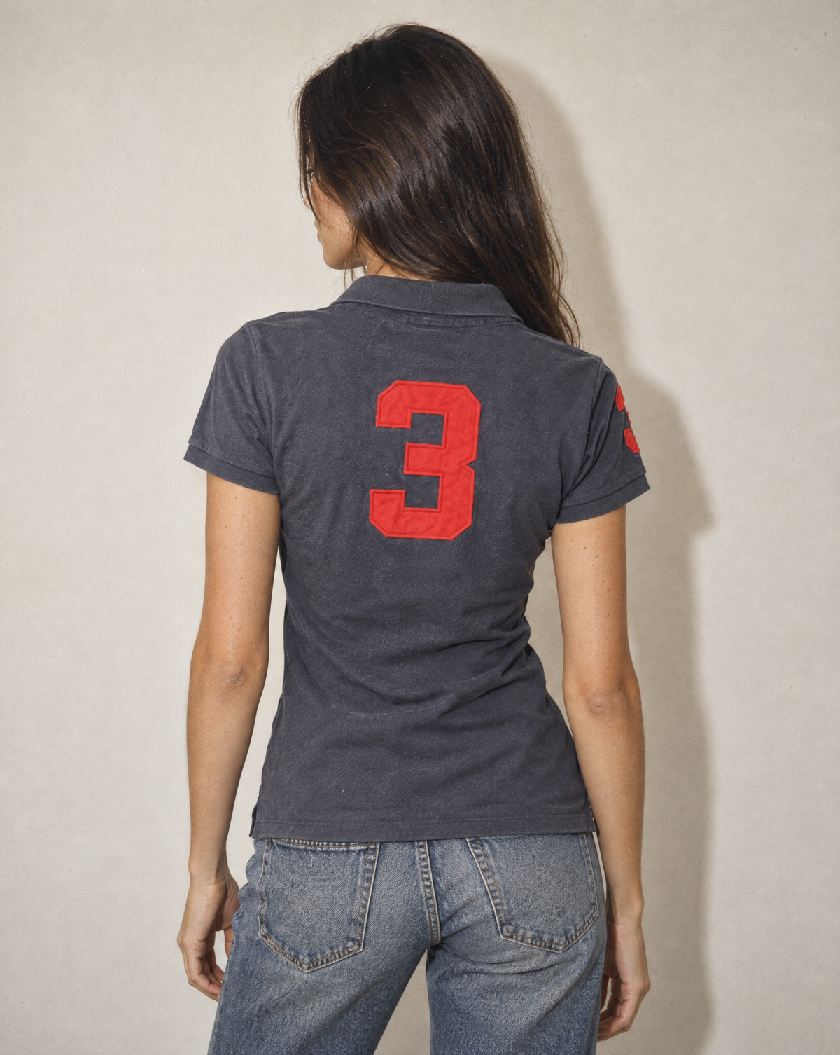 A woman viewed from behind wearing a dark charcoal gray short-sleeve polo shirt with a large red number 3 printed on the back, paired with blue jeans against a plain beige wall.
