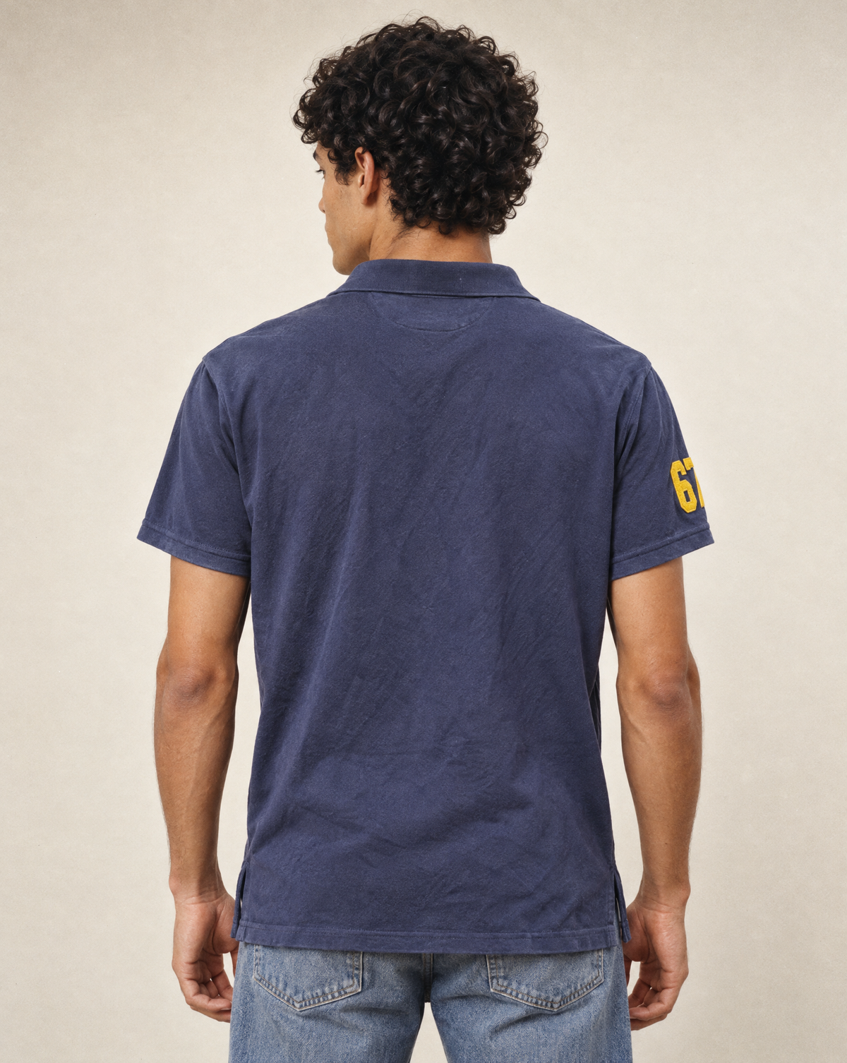 A man viewed from the back wearing a navy blue short-sleeve polo shirt with a yellow number 6 patch on the left sleeve, paired with blue jeans, against a neutral beige background.