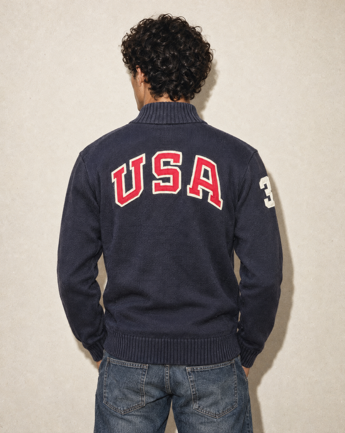 A man viewed from behind wearing a navy blue knit sweater with 'USA' in red and white block letters across the back and a white number 3 on the sleeve, paired with blue jeans against a textured beige wall.