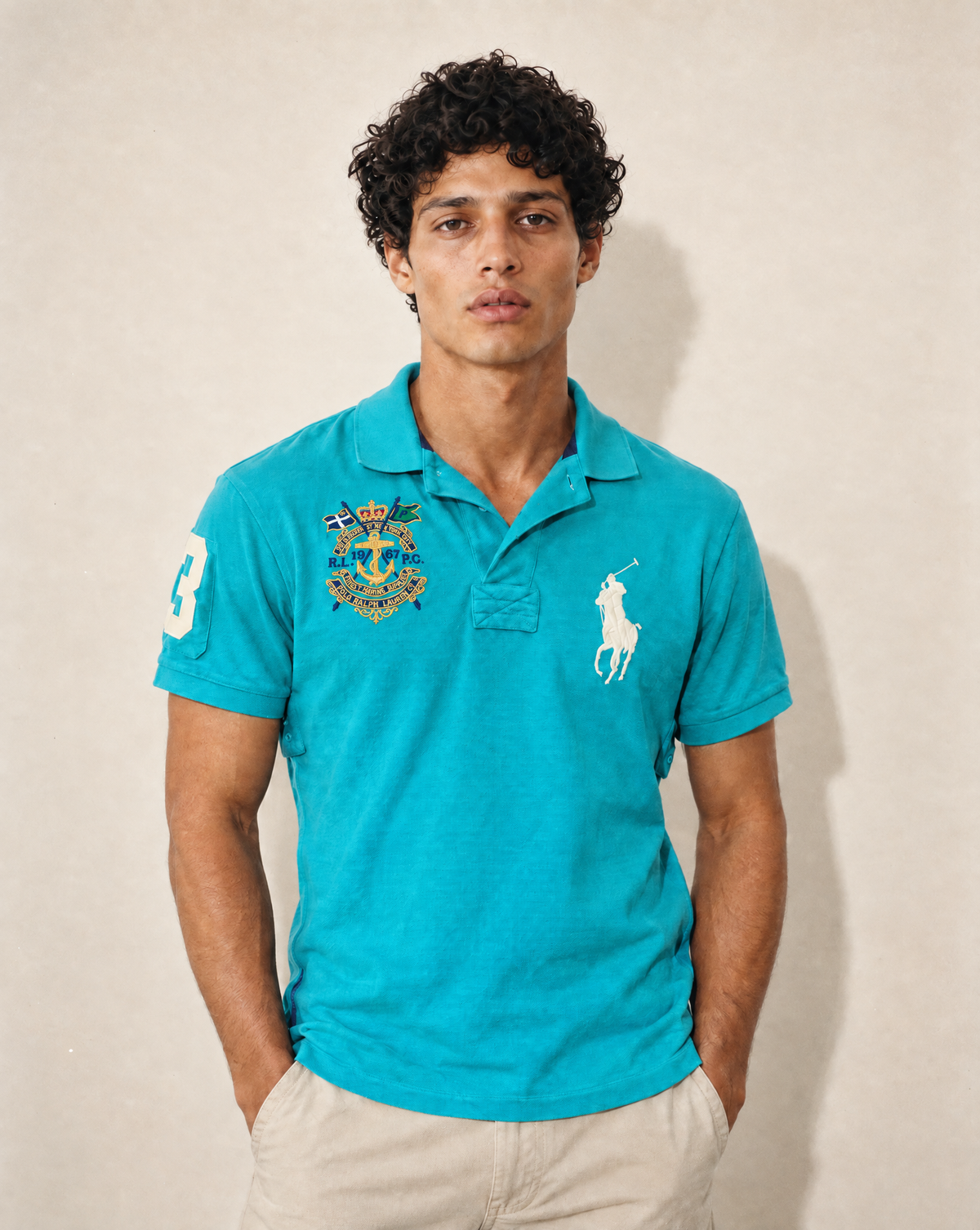 A man wearing a bright turquoise polo shirt with the white embroidered Polo Ralph Lauren logo on the left chest and a multicolor crest on the right chest, paired with beige trousers, standing against a textured beige wall.