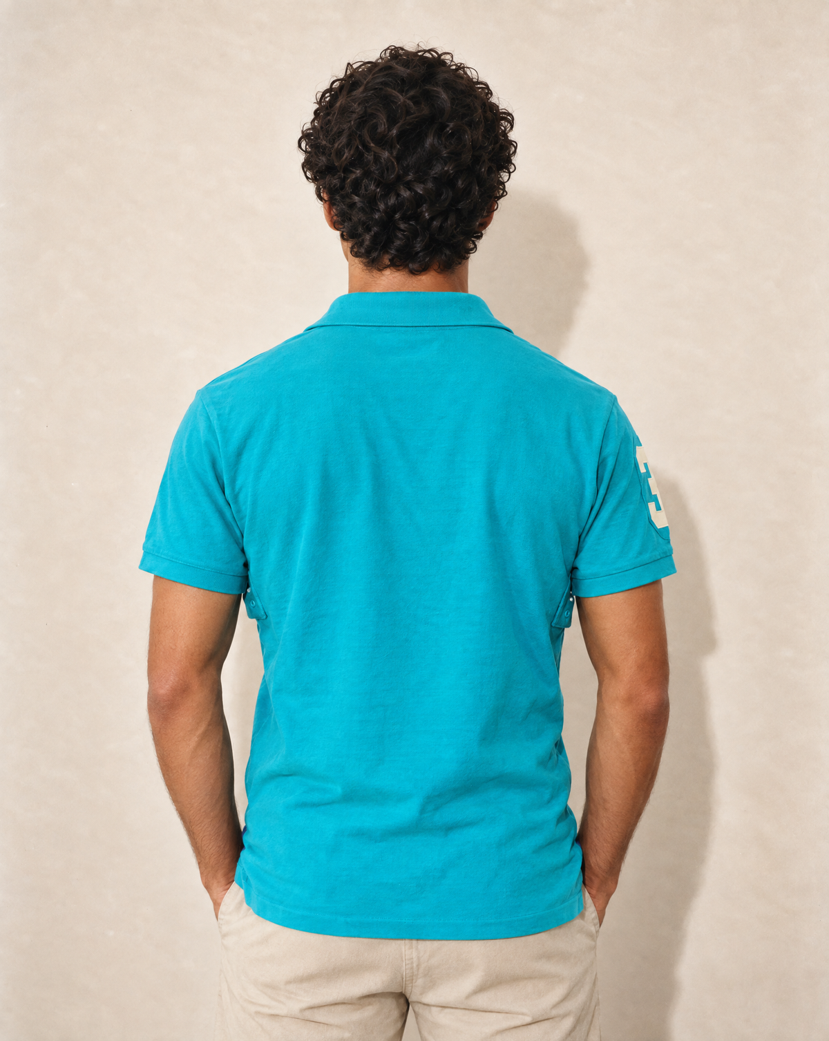 A man viewed from behind wearing a bright turquoise short-sleeve polo shirt and light beige pants, standing against a textured beige wall.