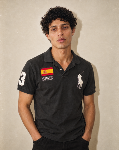 A man wearing a black Polo Ralph Lauren polo shirt with a Spanish flag graphic and 'SPAIN' text on the chest, posed against a textured beige wall.