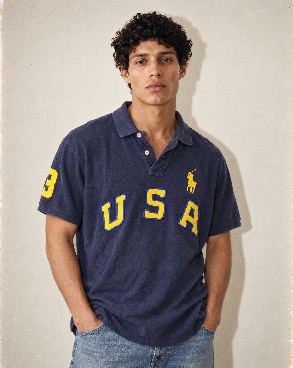 A man wearing a vintage-style navy blue polo shirt with yellow 'USA' lettering and the signature Polo Ralph Lauren pony logo, paired with blue jeans against a textured beige wall.