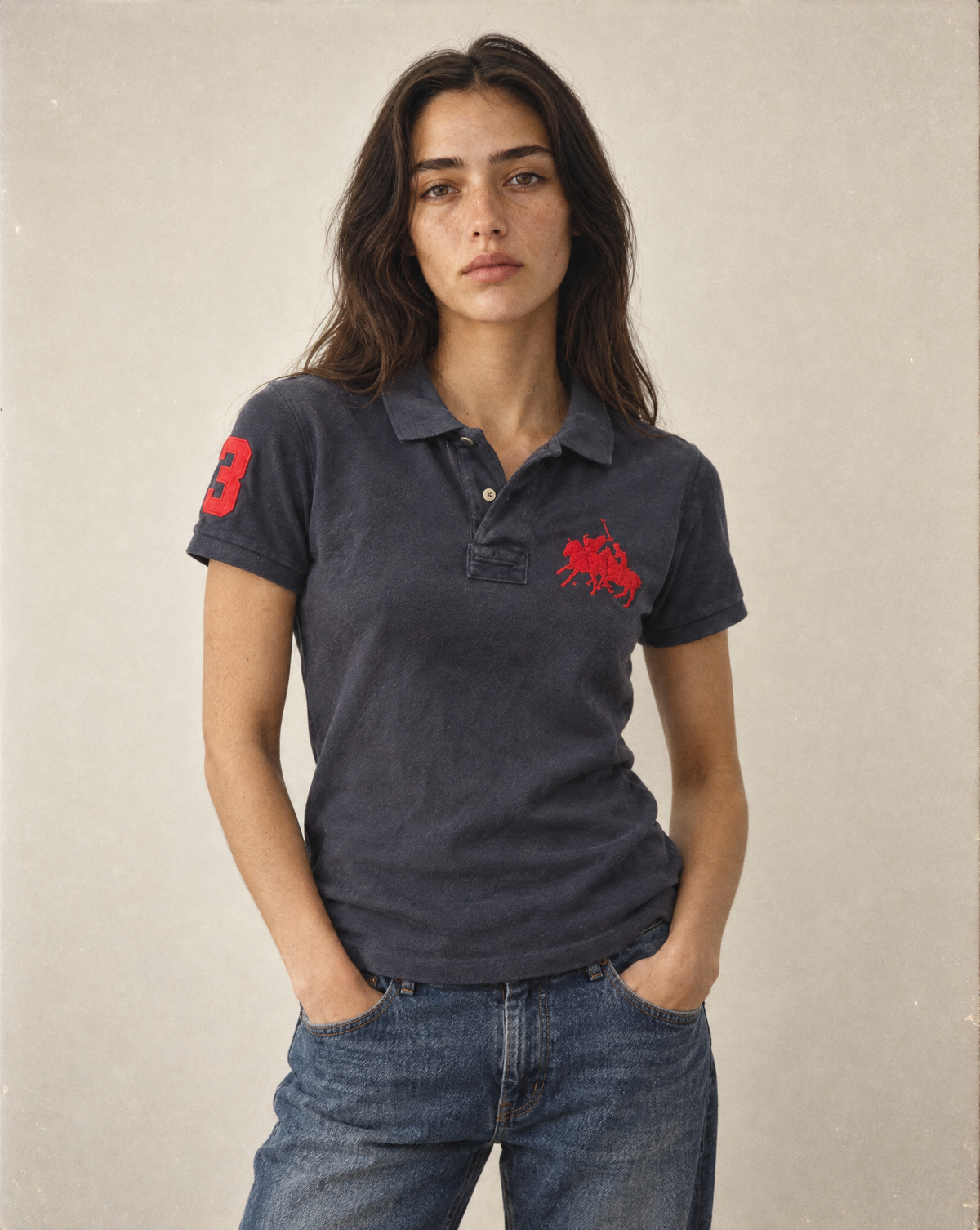A woman modeling a dark charcoal gray polo shirt with red embroidered graphics on the chest and sleeve, paired with blue jeans against a neutral beige background.