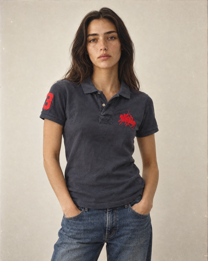 A woman modeling a dark charcoal gray polo shirt with red embroidered graphics on the chest and sleeve, paired with blue jeans against a neutral beige background.
