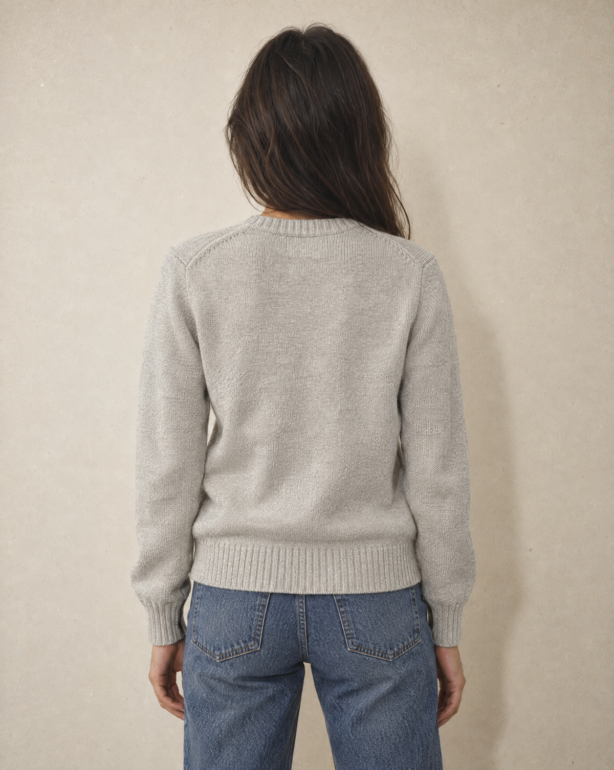 A woman viewed from behind wearing a light gray knit sweater and blue jeans, standing against a textured beige wall.