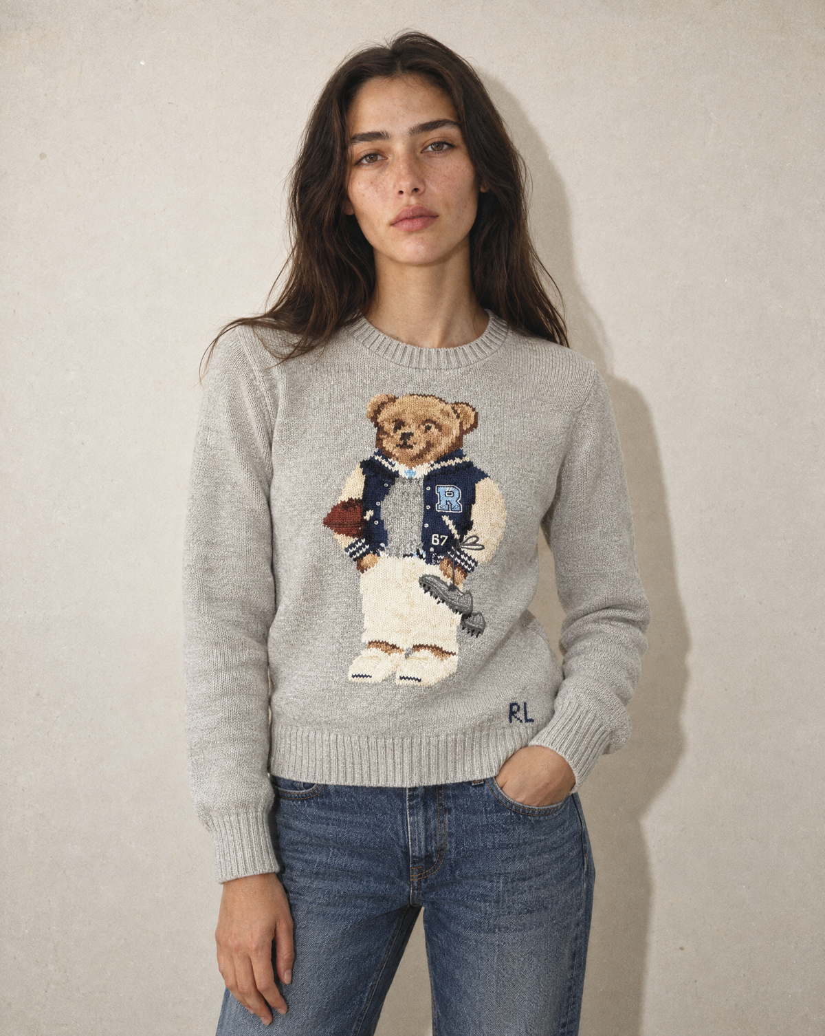 A woman wearing a light gray knit sweater with a large embroidered teddy bear graphic on the front, paired with blue jeans, standing against a textured beige wall.