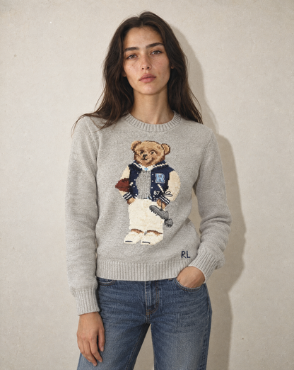 A woman wearing a light gray knit sweater with a large embroidered teddy bear graphic on the front, paired with blue jeans, standing against a textured beige wall.