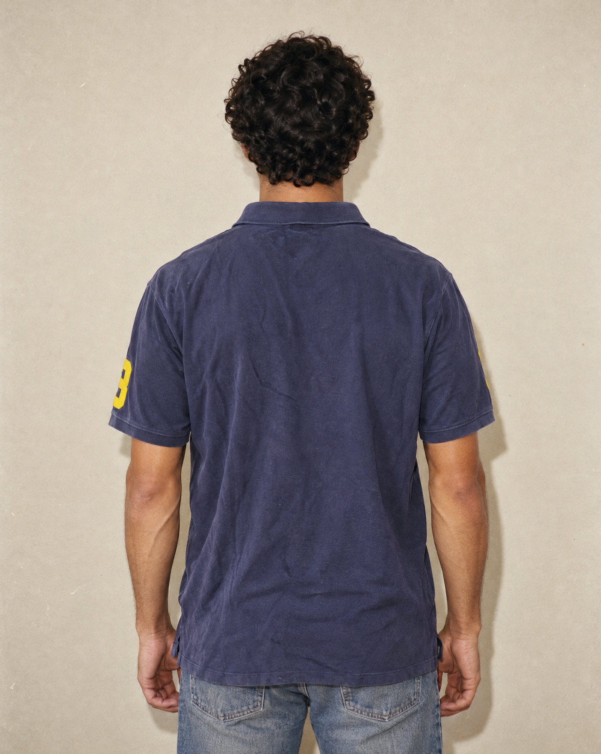 A man viewed from behind wearing a navy blue short-sleeve polo shirt with a yellow number 8 on the sleeve, paired with blue jeans, standing against a textured beige wall.