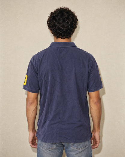 A man viewed from behind wearing a navy blue short-sleeve polo shirt with a yellow number 8 on the sleeve, paired with blue jeans, standing against a textured beige wall.